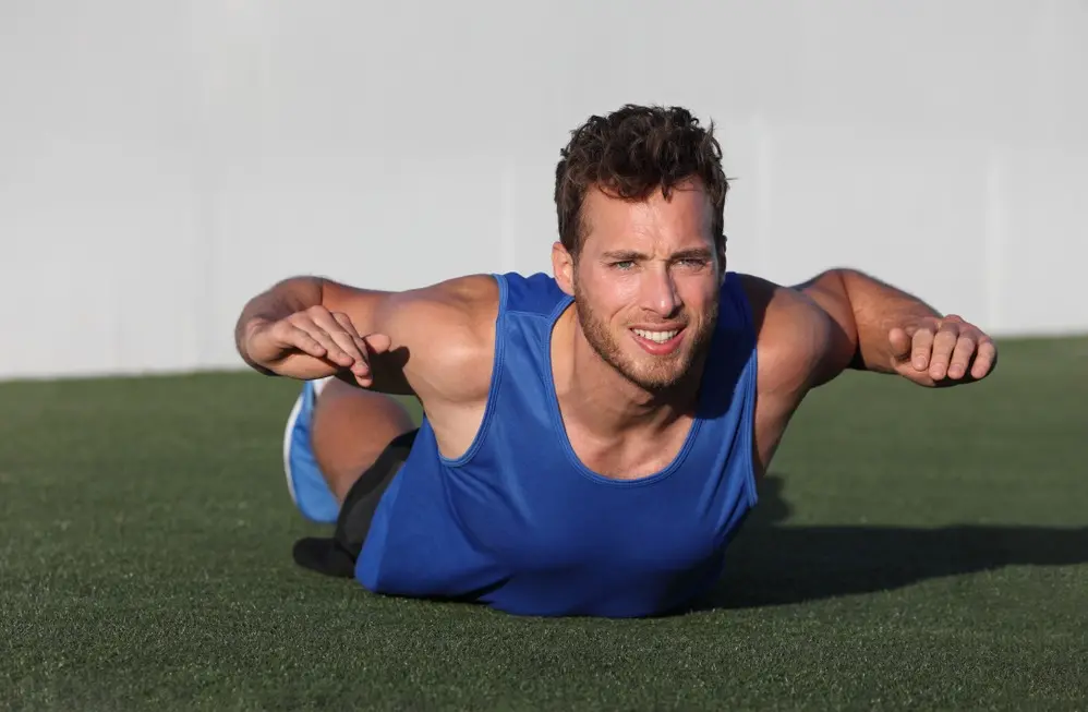 Homem branco e loiro, vestido com regata azul e short preto, faz o exercício superman no gramado.