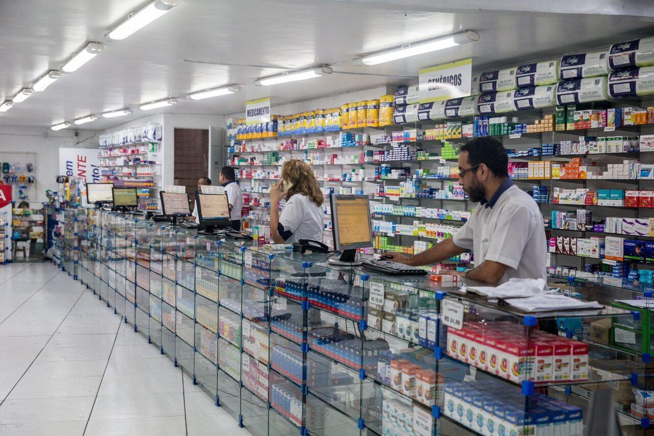 Farmácia com atendentes no balcão, prateleiras cheias de medicamentos e computadores.