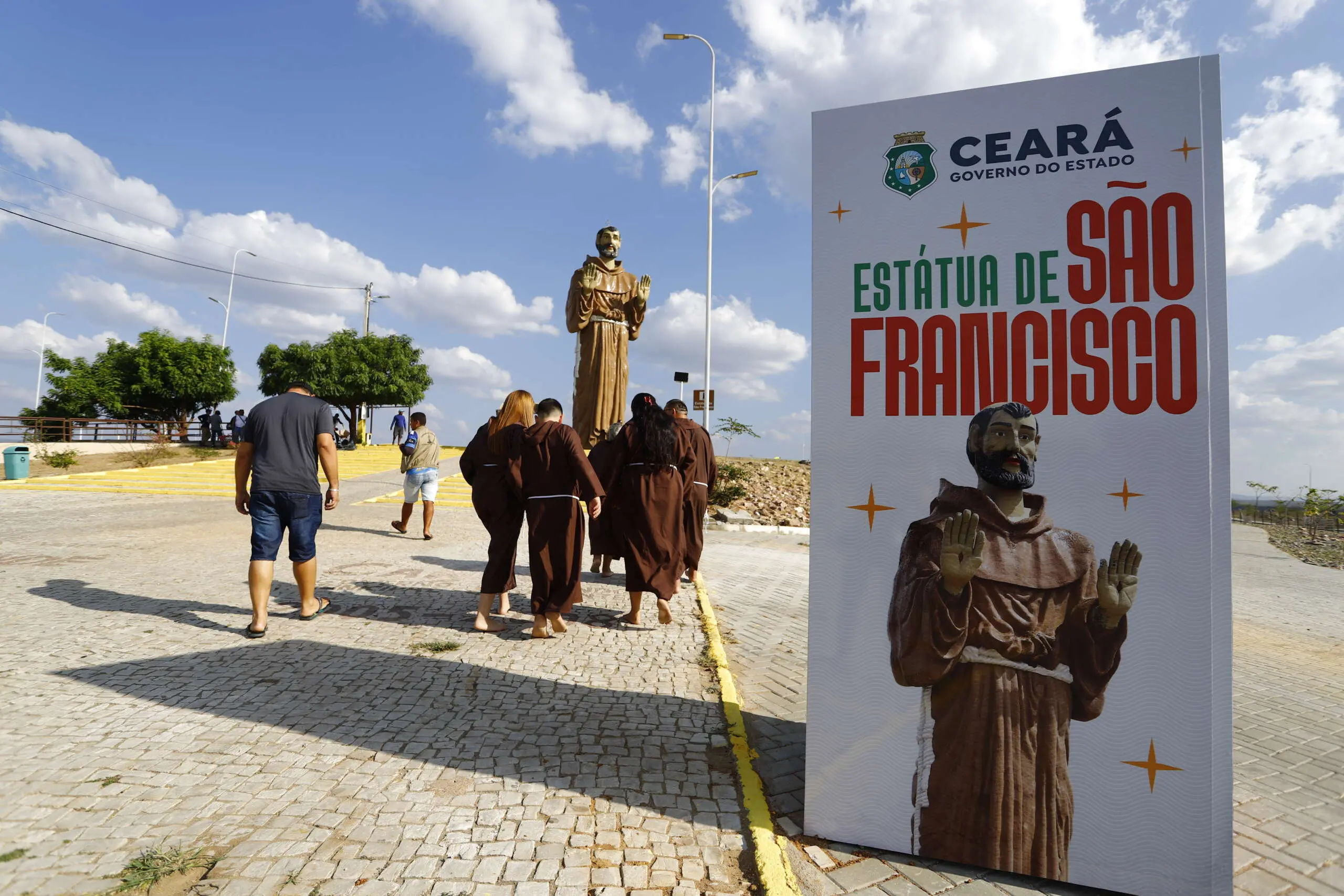 Imagem mostra, em primeiro plano, banner com nome Estátua de São Francisco e imagem do santo. Em segundo plano, homens vestidos de franciscanos caminham em direção à estátua gigante do santo, em Canindé, no Ceará.
