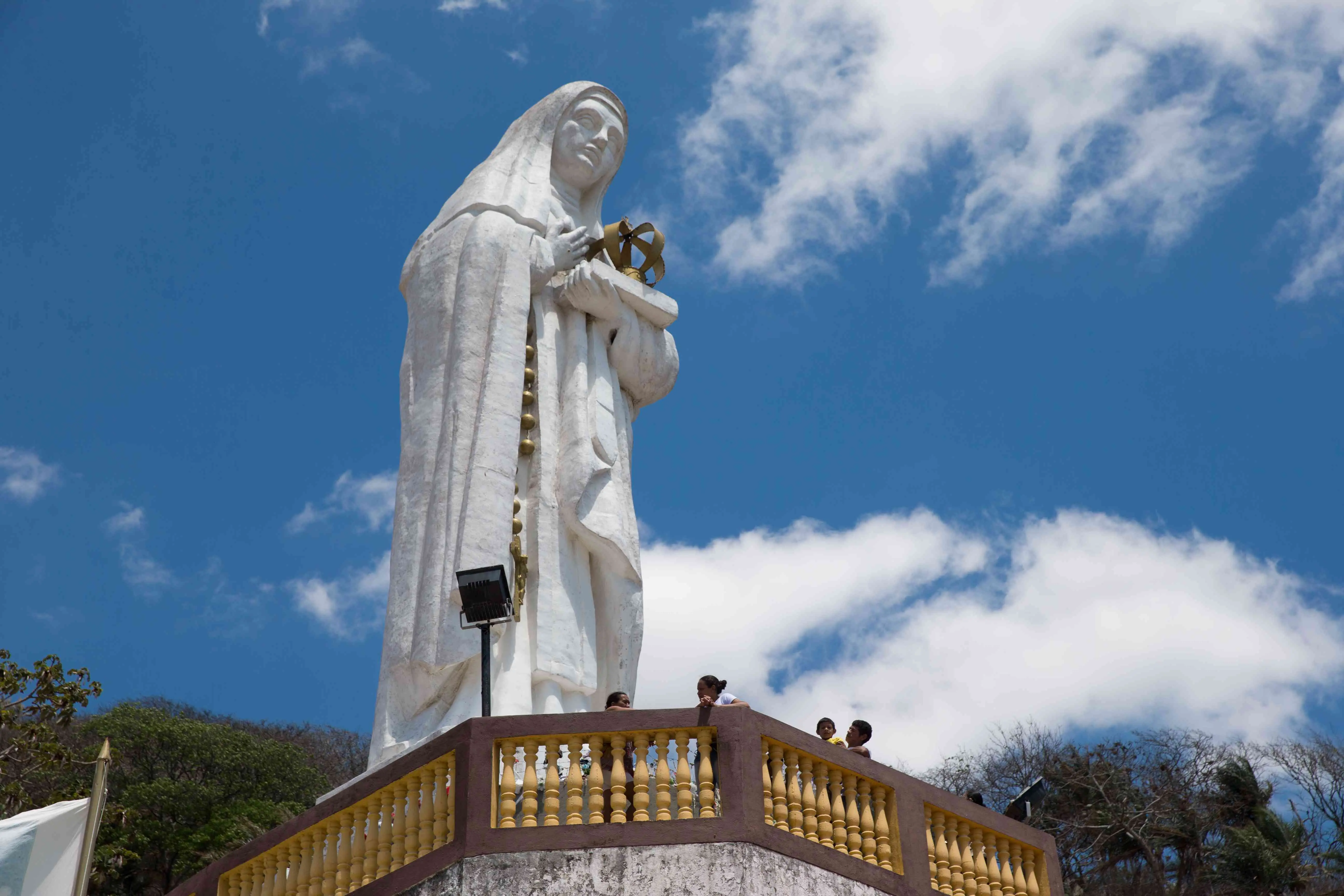 Imagem mostra estátua de Santa Edwiges, toda branca, cerca por estrutura de cobogós pintados em amarelo e marrom, onde se apoiam os fiéis. Atrás, o céu azul e nuvens brancas.