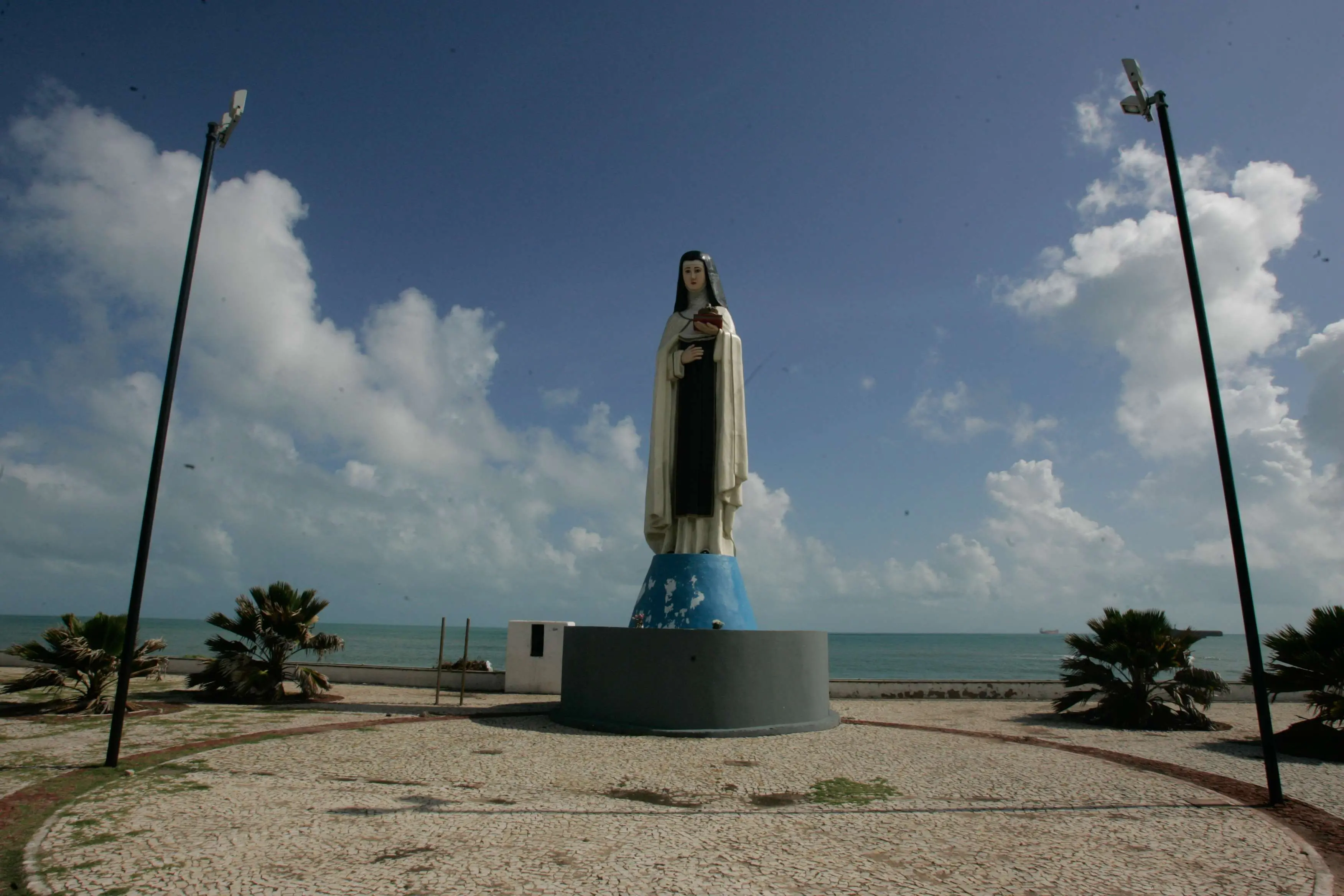 Imagem mostra estátua de Santa Edwiges na orla de Fortaleza. Atrás da santa, é possível ver o mar verde e um céu azul com nuvens brancas.