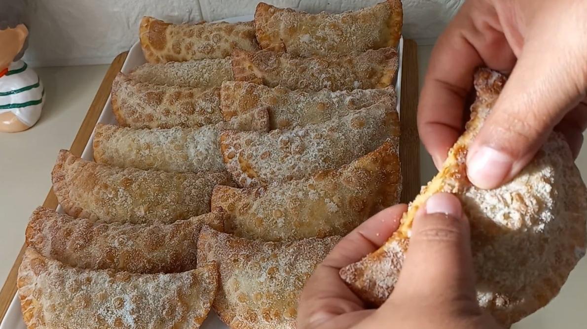 Uma travessa retangular branca cheia de pasteizinhos de festa típicos de Pernambuco, que são pequenos pastéis fritos com formato de meia-lua, cobertos com uma camada de açúcar e canela. À direita, uma mão segura um dos pastéis, mostrando seu recheio e a textura crocante da massa.