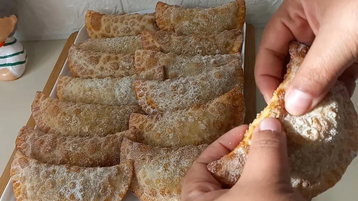 Uma travessa retangular branca cheia de pasteizinhos de festa típicos de Pernambuco, que são pequenos pastéis fritos com formato de meia-lua, cobertos com uma camada de açúcar e canela. À direita, uma mão segura um dos pastéis, mostrando seu recheio e a textura crocante da massa.