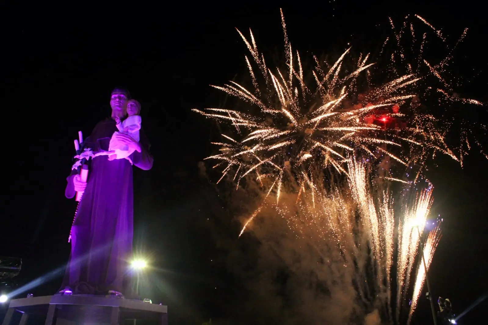 Imagem noturna mostra estátua de Santo Antonio em Barbalha, no Ceará. Ao lado dela, fogos de artifício.