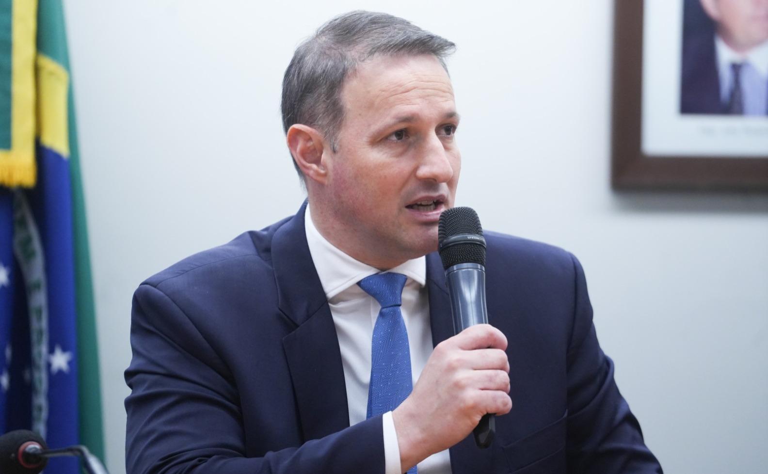 Guilherme Derrite é um homem branco de meia-idade e cabelo curto grisalho. Na foto, ele está de terno azul, camisa social branca e gravata azul, segurando microfone. Imagem usada em matérias sobre os principais pontos do projeto Antifacção.