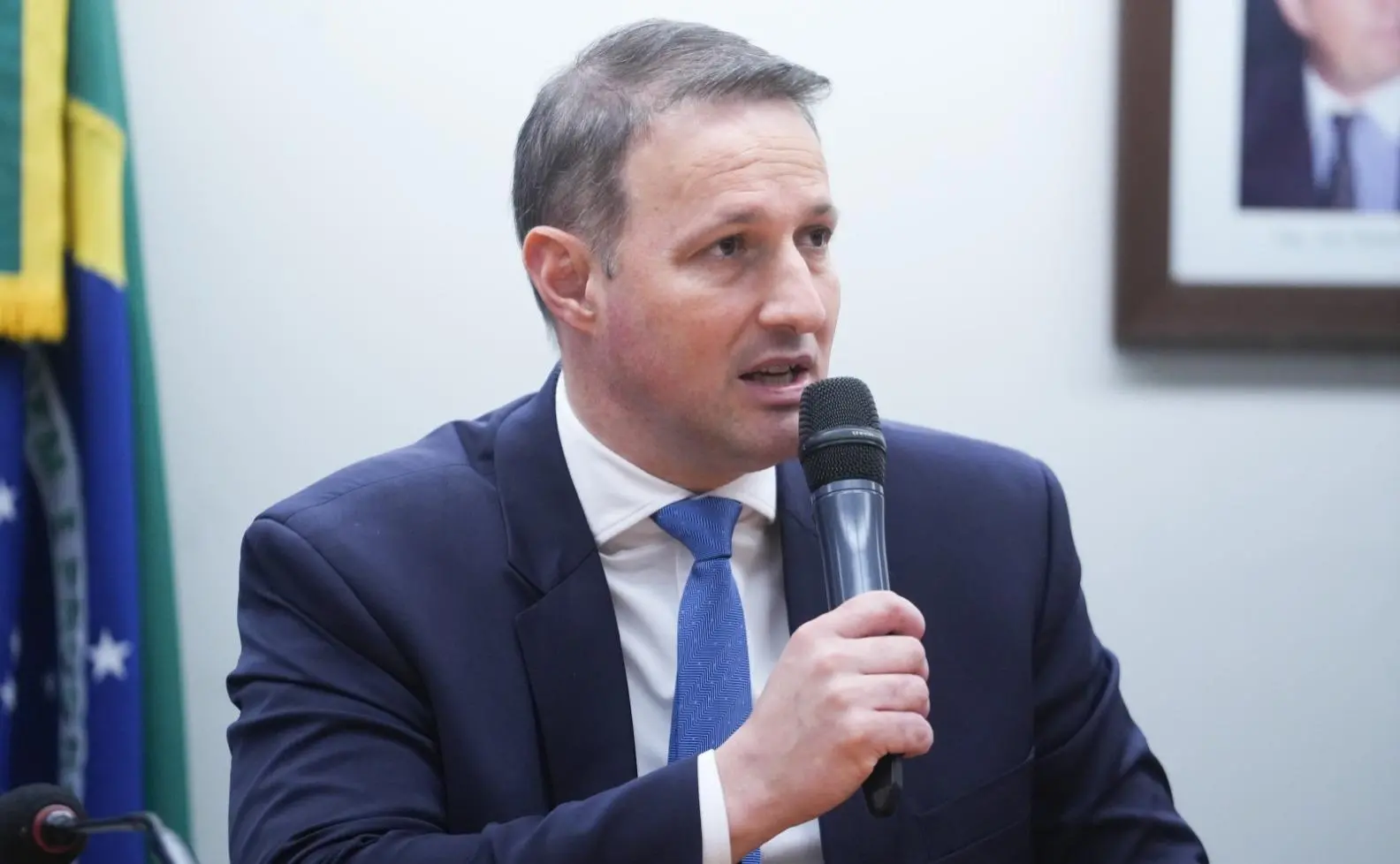 Guilherme Derrite é um homem branco de meia-idade e cabelo curto grisalho. Na foto, ele está de terno azul, camisa social branca e gravata azul, segurando microfone. Imagem usada em matérias sobre os principais pontos do projeto Antifacção.