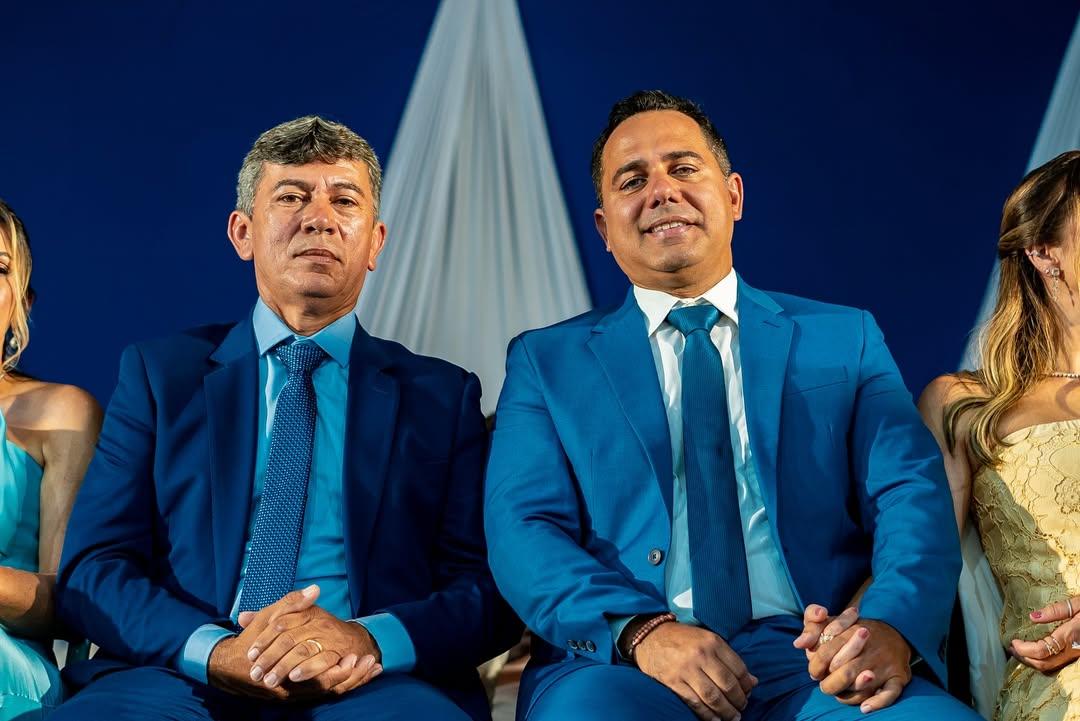 O vice-prefeito de Iguatu Francisco Frutas, a direita, e o prefeito de Iguatu Roberto Filho, a esquerda. Eles sentados lado a lado usam ternos azuis e gravatas em tons de azul, posando para foto durante evento formal com fundo azul e tecido branco decorativo ao centro.