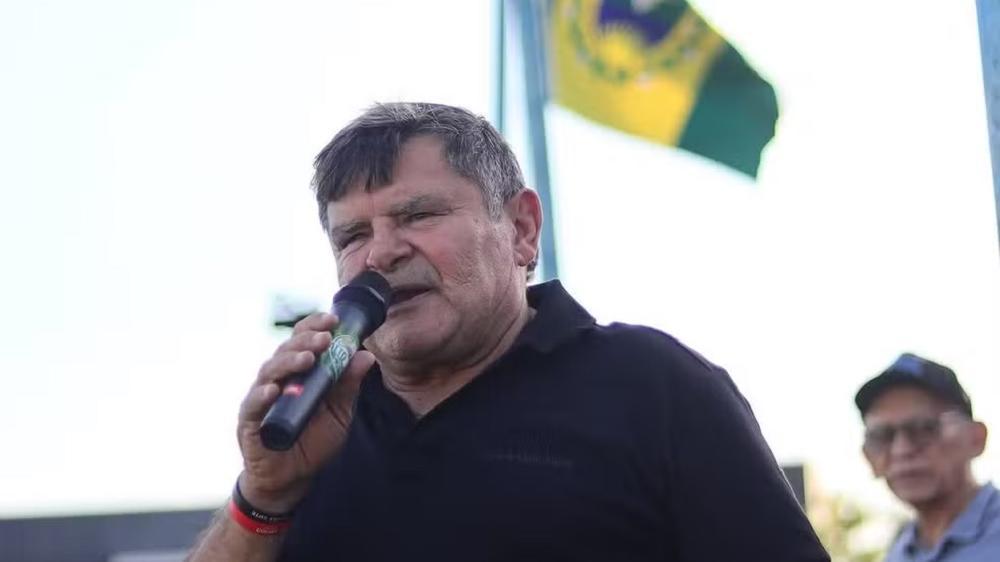 Homem de meia-idade fala ao microfone durante um evento ao ar livre, vestindo camisa preta. Ao fundo, há uma bandeira do Brasil parcialmente visível e outra pessoa desfocada. O céu está claro, indicando que a foto foi tirada durante o dia.