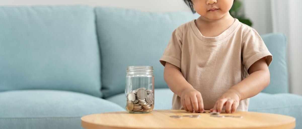 Na foto, uma criança mexe com moedas que ela retirou de um cofre de vidro. Ao fundo, há um estofado azul.