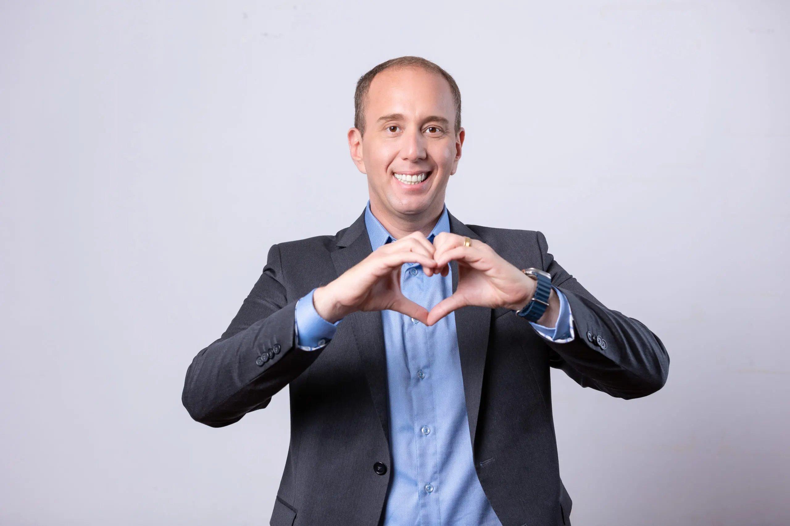 Imagem mostra Rodrigo Manga, prefeito de Sorocaba (SP) recém-afastado do cargo, sorridente com blazer cinza e camisa azul, formando um coração com as mãos em fundo claro.