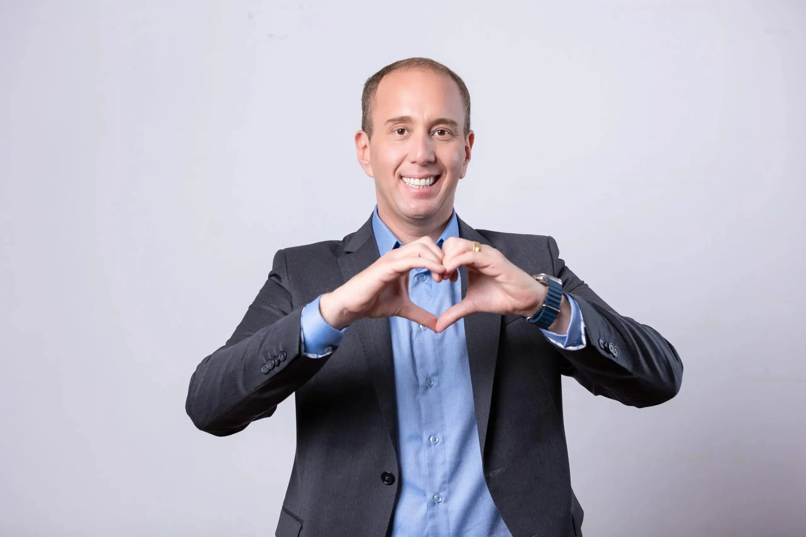 Imagem mostra Rodrigo Manga, prefeito de Sorocaba (SP) recém-afastado do cargo, sorridente com blazer cinza e camisa azul, formando um coração com as mãos em fundo claro.