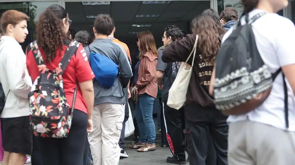 Grupo de jovens, muitos com mochilas, reunidos na entrada de um edifício, provavelmente uma escola ou universidade.