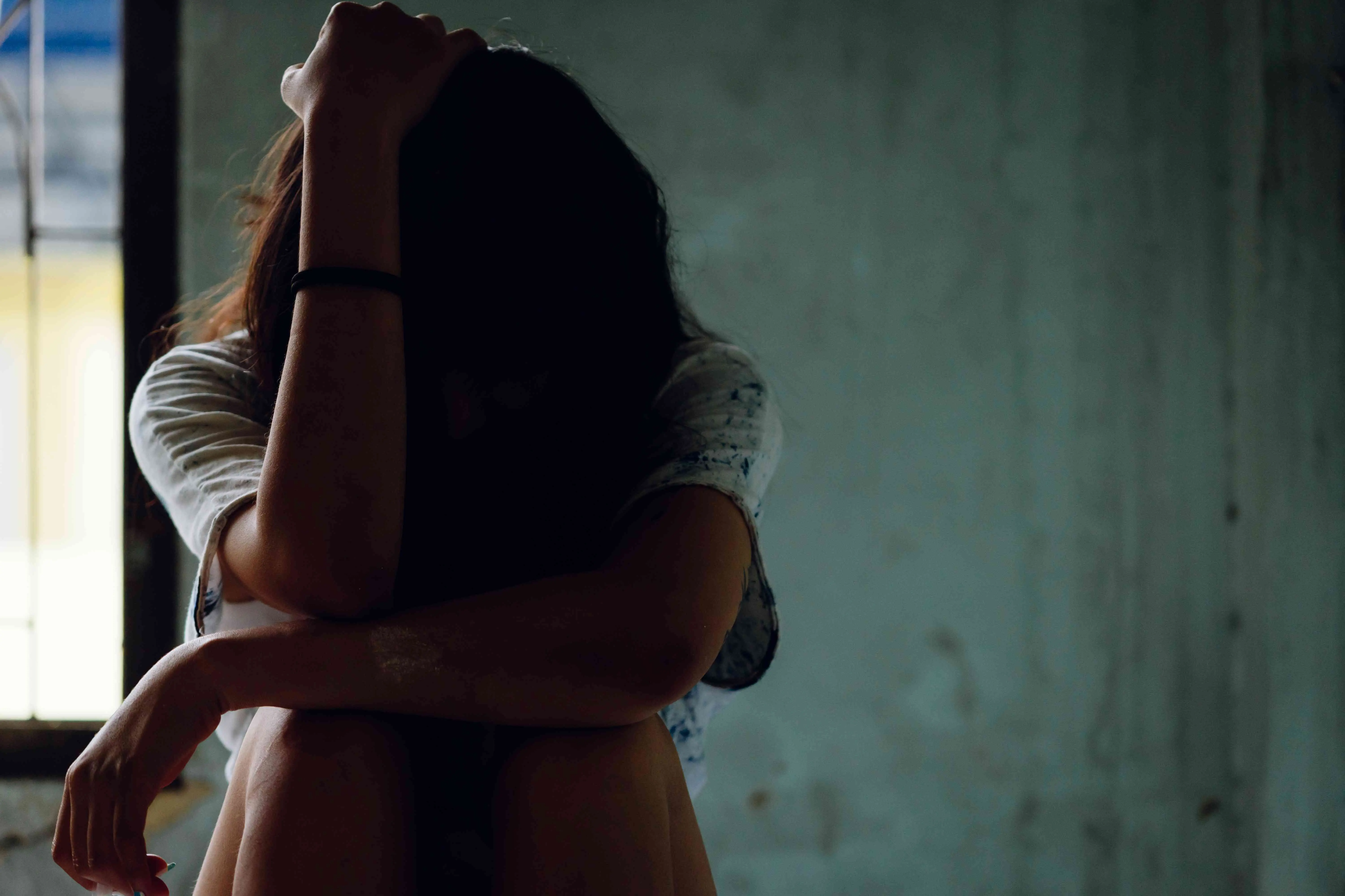 Imagem mostra silhueta de adolescente sentada em um canto escuro, com a cabeça entre os braços, transmitindo angústia e vulnerabilidade, ilustrando abuso sexual de vunerável.