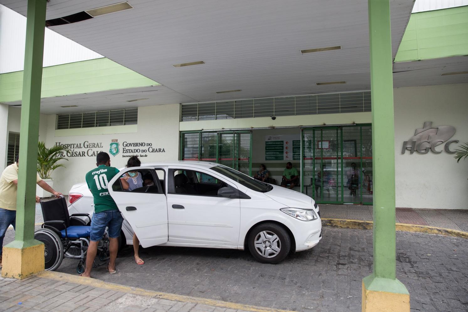 Fachada do Hospital Dr. César Cals, pintada de verde e com logotipo do Governo do Ceará. Há um carro branco na frente. Um homem ajuda uma mulher de máscara a sentar em uma cadeira de rodas.