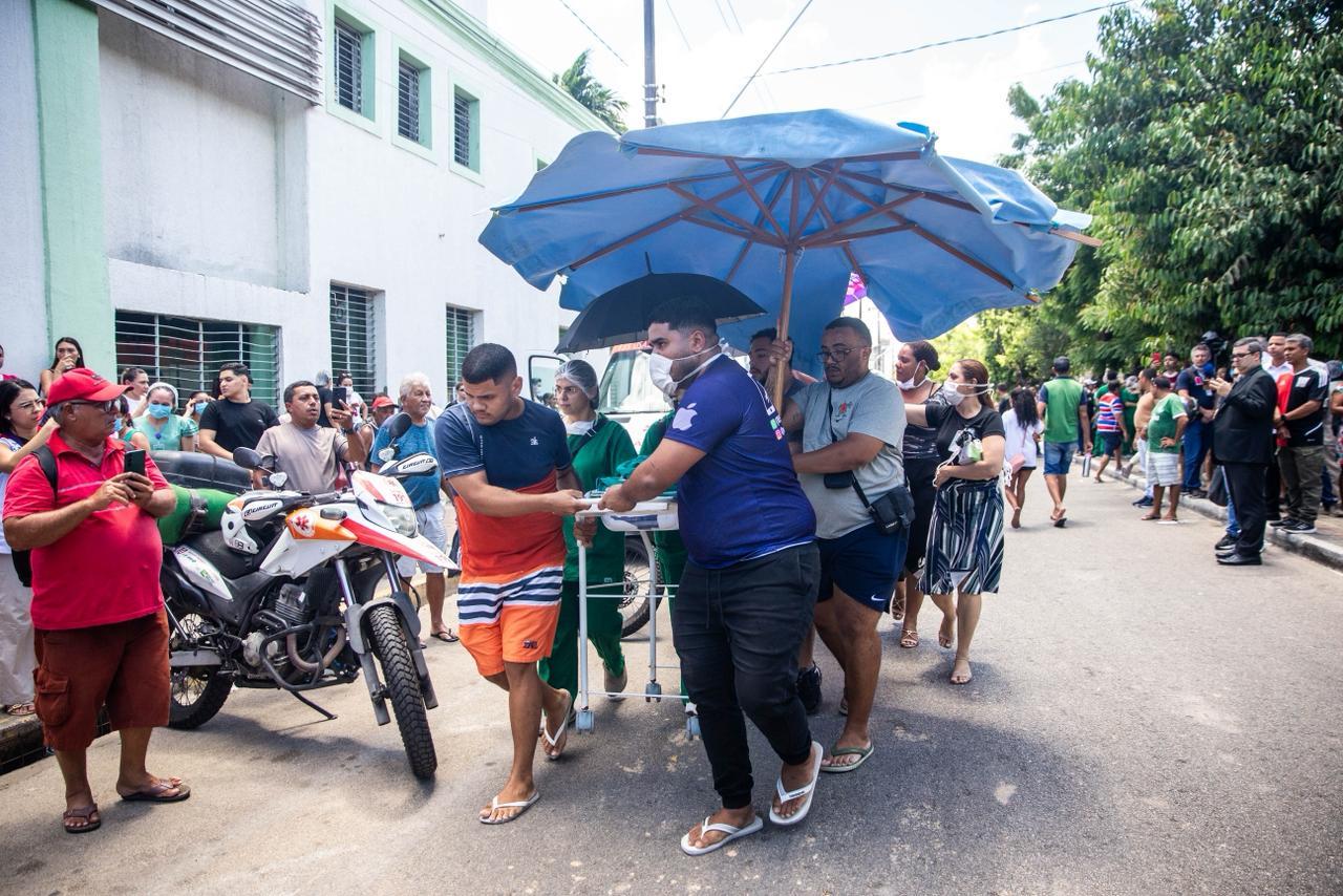 Foto de uma multidão em uma rua sob sol forte, transportando uma maca improvisada com alguém deitado, que é protegido por um grande guarda-sol azul-claro. O grupo de apoio, incluindo pessoas de uniforme de saúde, acompanha a maca para cima de um edifício branco.