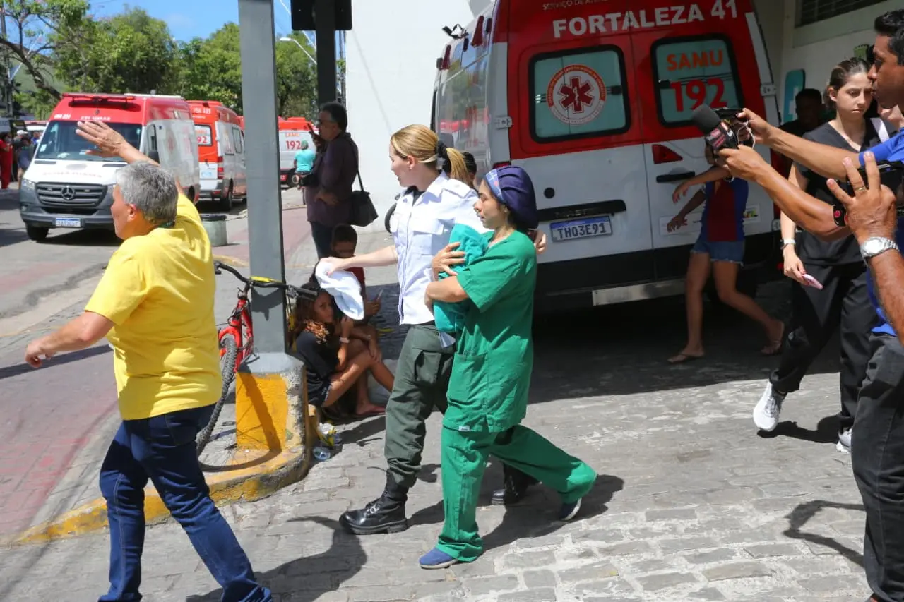 Uma profissional de saúde, vestindo uniforme cirúrgico verde, carrega um bebê ou criança pequena enrolado(a) em seus braços, enquanto caminha rapidamente. Ela está de perfil e de cabelo preso com um gorro azul. Ao seu lado, uma outra profissional de saúde, de uniforme branco e calças escuras, acompanha o passo. Ao fundo, há ambulâncias vermelhas e brancas do SAMU (Serviço de Atendimento Móvel de Urgência) com os números 
