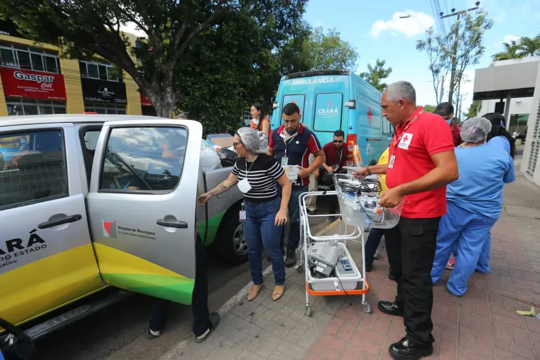 Uma equipe de profissionais, alguns vestindo roupas civis e outros uniformes de saúde (azul claro), está engajada no transporte de equipamentos médicos. Um homem de camisa vermelha está puxando um carrinho com rodízios contendo monitores e outros aparelhos médicos hospitalares. Ao seu lado, uma mulher de camisa listrada preta e branca e calça jeans está carregando itens ao lado de uma caminhonete prateada com o logo do 