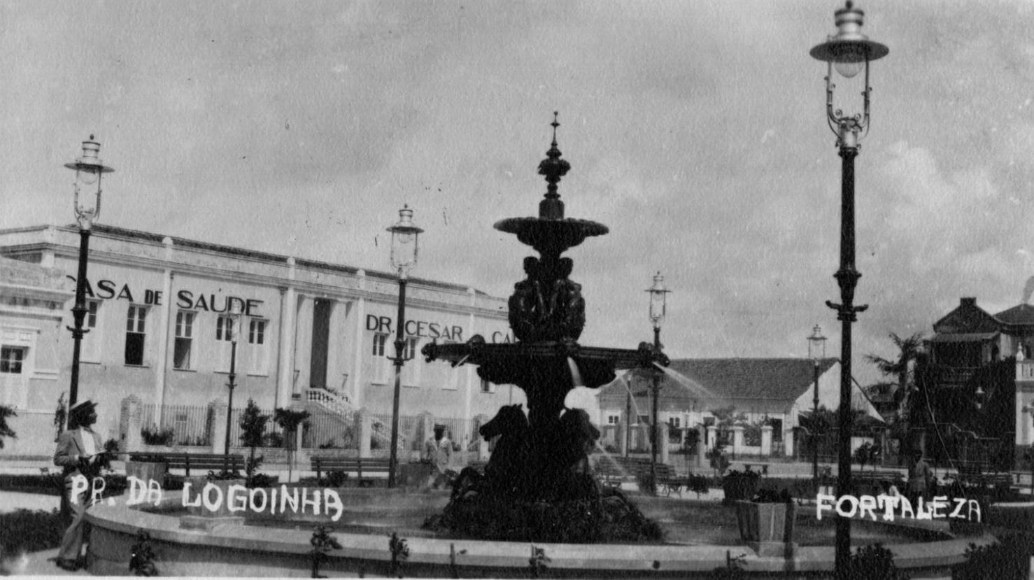 Foto em preto e branco mostra uma praça pública em Fortaleza, com uma fonte de água ao centro. Ao redor dela, há postes de lamparinas e alguns bancos. Ao fundo, há um prédio extenso com o letreiro Casa de Saúde Dr. César Cals.