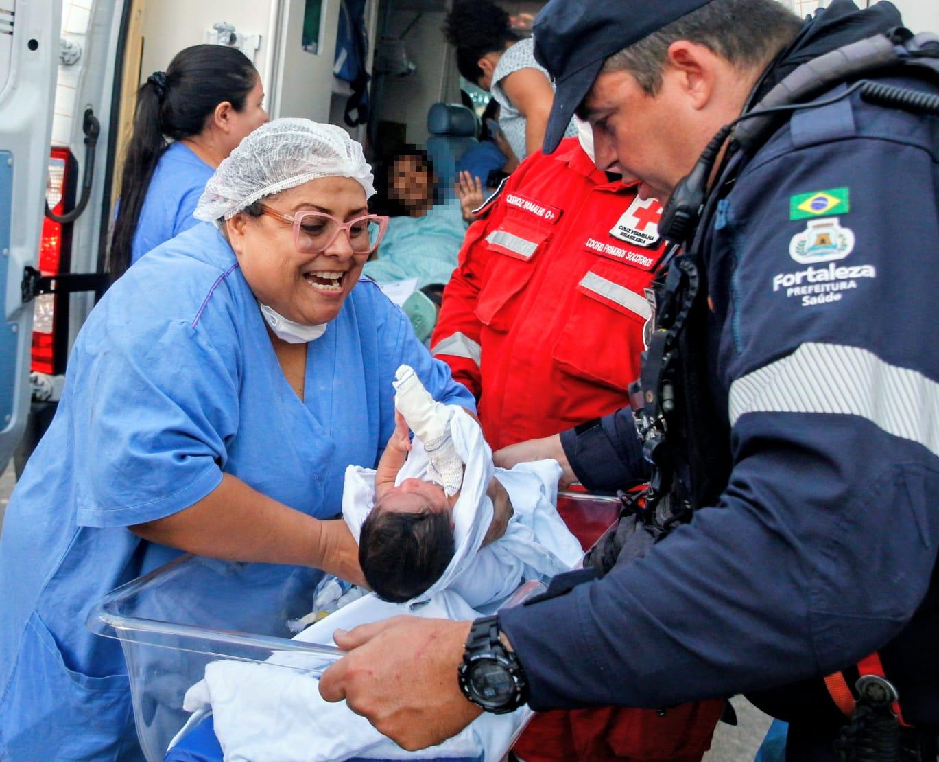 Profissionais de saúde trabalham juntos para transportar um recém-nascido envolto em mantas, enquanto uma mulher em maca dentro da ambulância observa. A cena ocorre ao lado de um veículo de emergência, transmitindo urgência e cuidado.