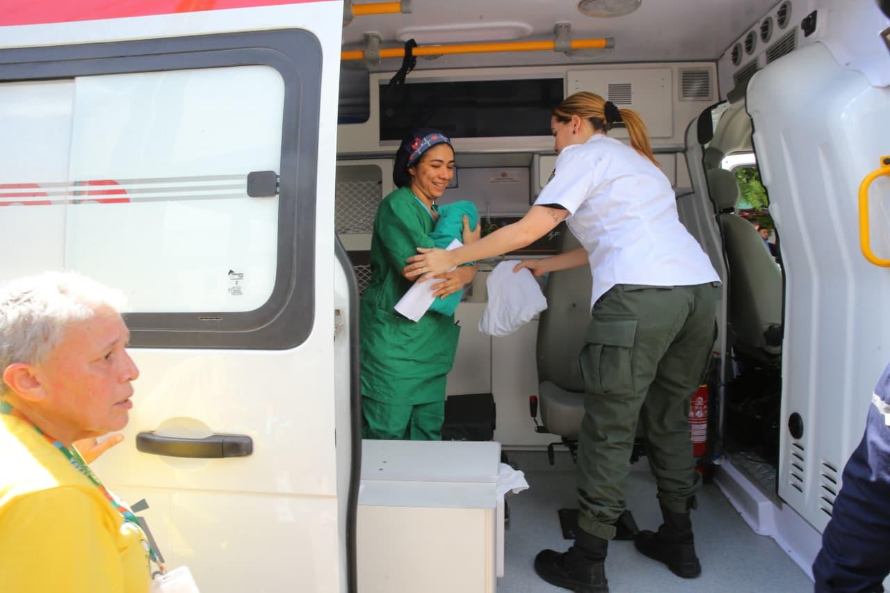 Dentro de uma ambulância, uma profissional de saúde vestindo roupa cirúrgica verde segura um bebê recém-nascido envolto em um pano, enquanto outra profissional, de uniforme branco e calça verde, entrega um lençol. À frente, do lado de fora do veículo, uma pessoa de camiseta amarela observa a cena.