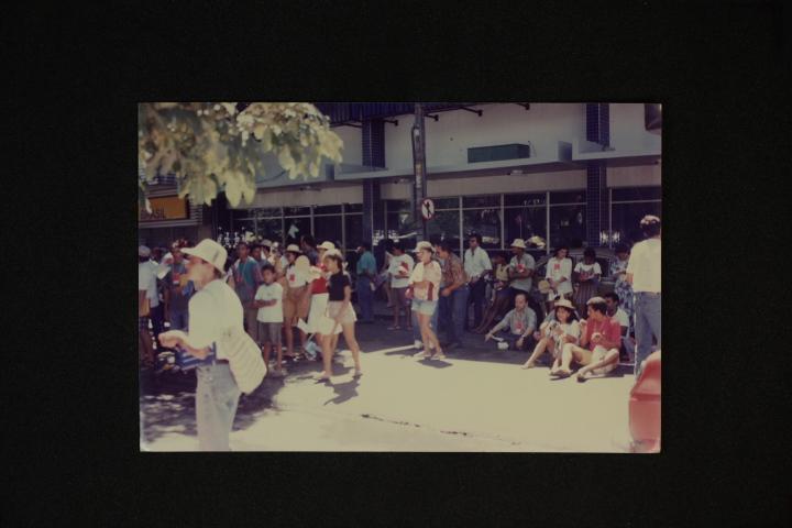 Foto antiga mostrando um grupo diverso de pessoas reunidas em uma rua e calçada ensolarada. Algumas estão em pé e outras sentadas no chão em frente a um prédio comercial moderno. Galhos de árvore sombreiam o canto superior esquerdo.