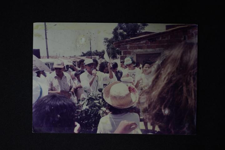 Foto antiga, desbotada, de um grupo de pessoas em um evento de rua. Um homem de boné claro no centro fala em um microfone para a multidão. Em primeiro plano, uma pessoa de chapéu de palha está de costas.