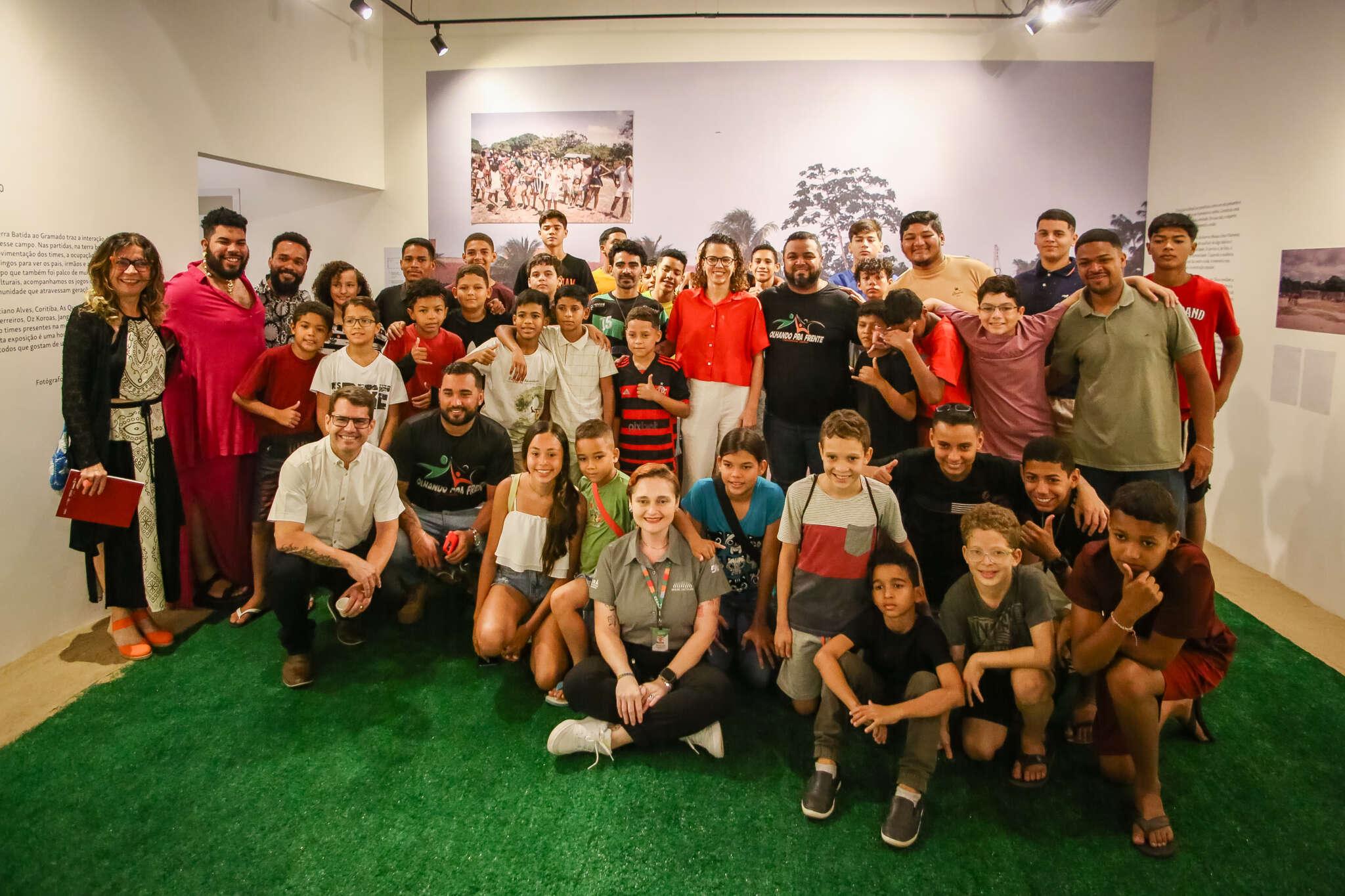 Foto de grupo grande de crianças, adolescentes e adultos, reunidos em uma sala de museu com piso de grama sintética. 