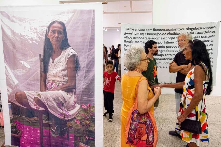 Fotografia em um museu. À esquerda, um grande banner exibe o retrato de uma mulher sentada. À direita, um pequeno grupo de visitantes (incluindo uma mulher de vestido laranja e uma de vestido estampado colorido) interage em frente a um painel de texto.