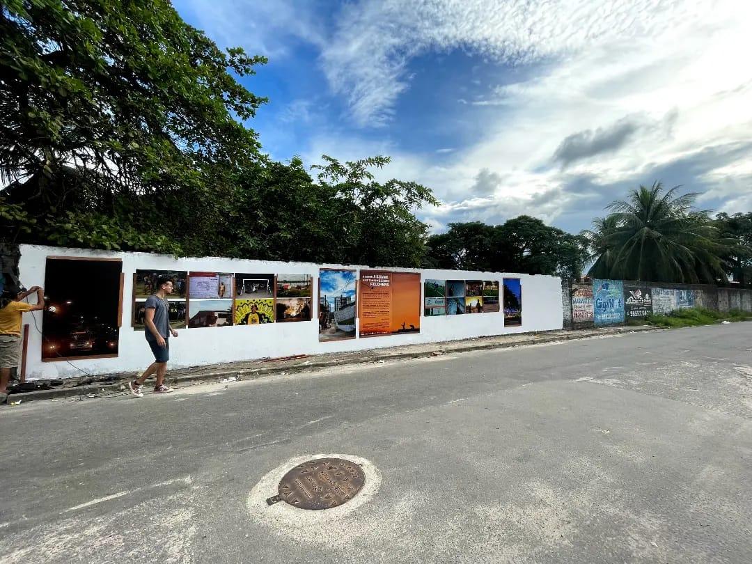 Fotografia de uma exposição de arte ao ar livre composta por painéis fotográficos e textuais em um muro branco baixo, na beira de uma rua asfaltada. Um jovem caminha em frente aos painéis. O céu é azul com nuvens.