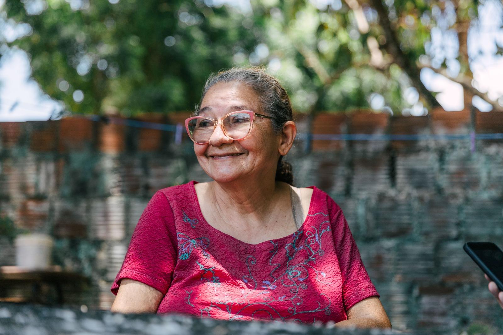 Retrato em plano médio de uma mulher de óculos rosa e camisa rosa/magenta, sorrindo amplamente e olhando para a esquerda. O fundo desfocado é uma parede de tijolos e árvores. Uma mão segurando um celular é visível no canto inferior direito.