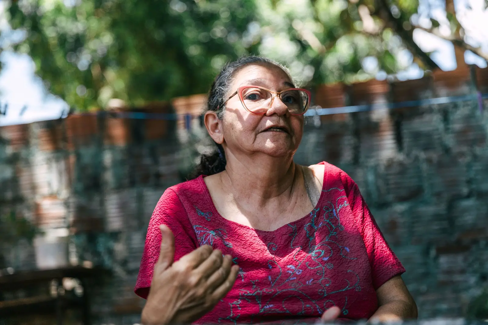 Retrato em plano médio de uma mulher de óculos rosa e camisa rosa/magenta, gesticulando com a mão direita, como se estivesse falando. O fundo desfocado é uma parede de tijolos e árvores.