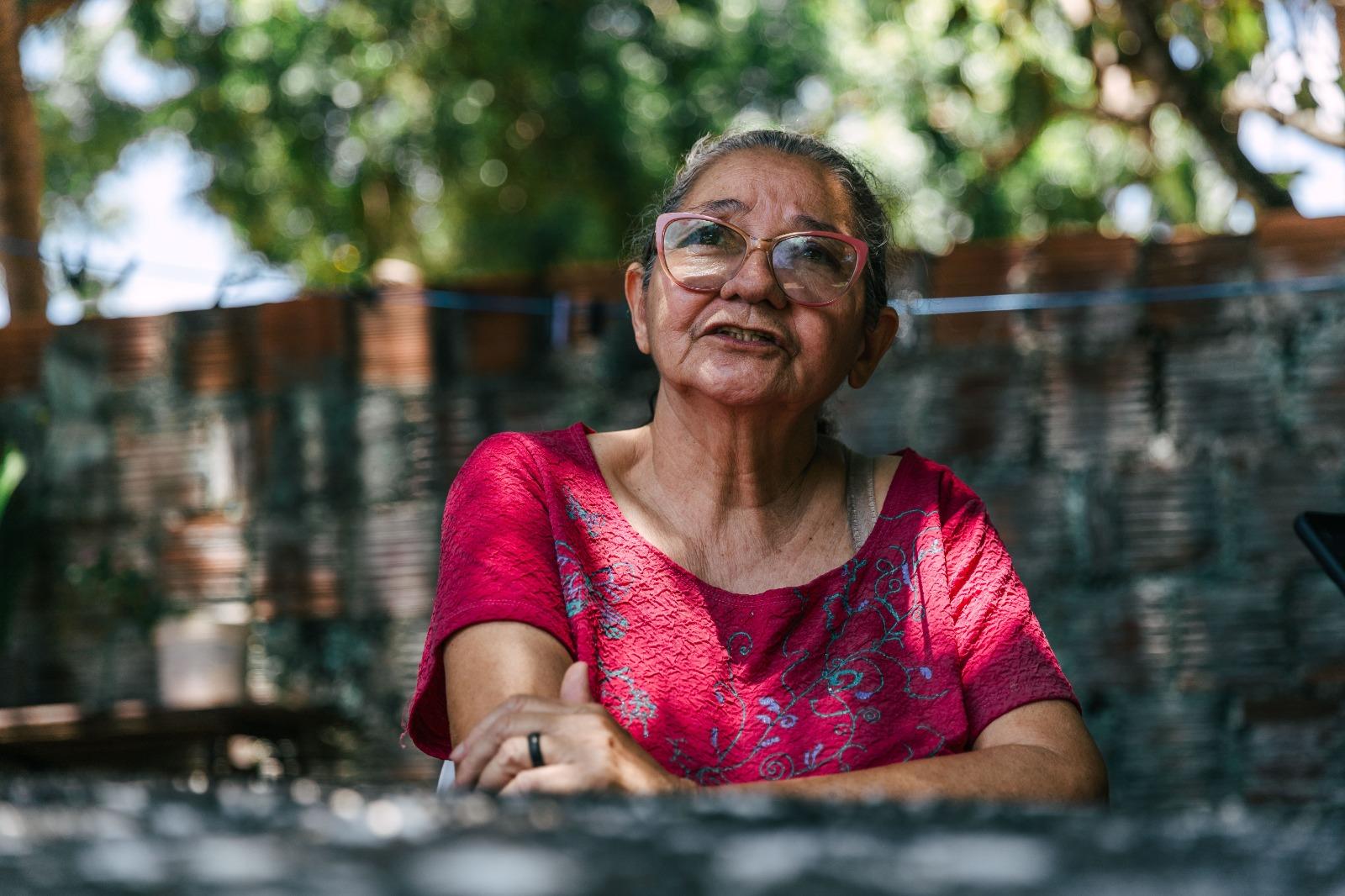 Retrato em plano médio de uma mulher de óculos rosa e camisa rosa/magenta, com os braços cruzados, sentada em ambiente externo. O fundo desfocado mostra uma parede de tijolos e árvores.