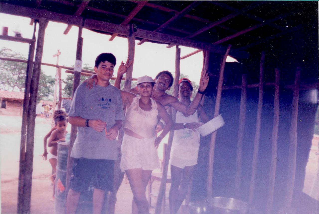 Foto antiga, escura, que mostra quatro adultos (três homens e uma mulher de boné) sorrindo e abraçados sob uma estrutura de madeira rústica e sombreada. Um deles levanta o braço.