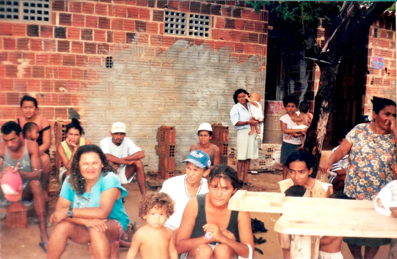 Foto antiga de um grupo de pessoas, incluindo adultos e bebês, posando para a câmera. O fundo é uma parede rústica de tijolos aparentes e reboco cinza.