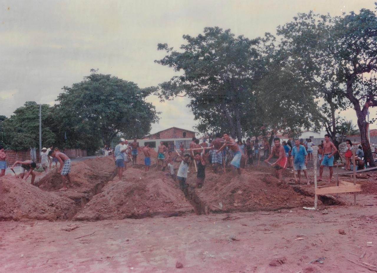 Foto antiga, com tons rosados, de um mutirão de homens cavando e movendo grandes montes de terra vermelha. Eles usam pás e enxadas. Grandes árvores e casas simples estão ao fundo.