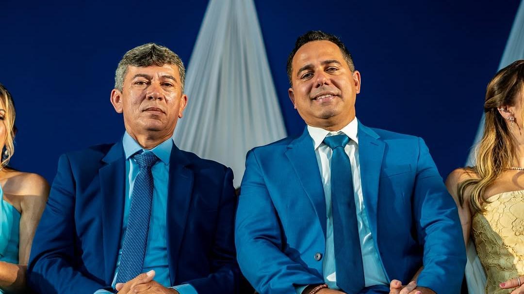 O vice-prefeito de Iguatu Francisco Frutas, a direita, e o prefeito de Iguatu Roberto Filho, a esquerda. Eles sentados lado a lado usam ternos azuis e gravatas em tons de azul, posando para foto durante evento formal com fundo azul e tecido branco decorativo ao centro.