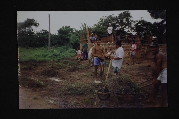 Foto antiga e escura de um mutirão de construção. Homens trabalham em um terreno de terra escura, usando carrinhos de mão para mover mato e vegetação. Ao fundo, uma estrutura inacabada de tijolos com pessoas observando.