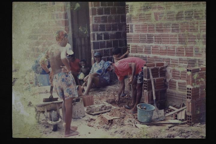 Foto antiga, manchada, de um mutirão de construção. O registro mostra homens e mulheres trabalhando com tijolos, materiais de construção e terra na frente de uma parede de tijolos aparentes.
