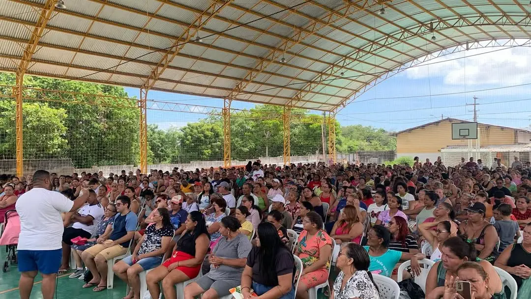 Foto de uma grande reunião comunitária em uma quadra coberta com teto de metal. Centenas de pessoas estão sentadas em cadeiras olhando para um orador (de costas, de branco) na extrema esquerda.