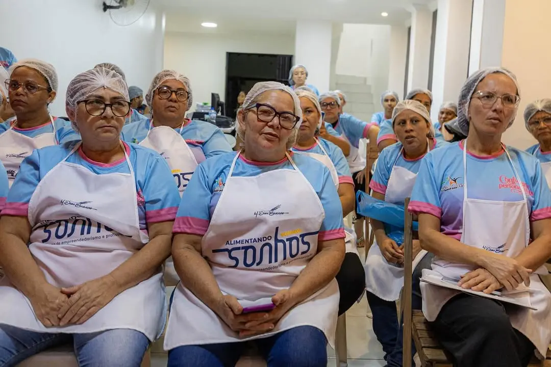 Retrato em plano médio de um grupo de mulheres adultas e de meia-idade sentadas em fileiras, participando de um curso. Elas usam aventais brancos e toucas de rede nos cabelos. O ambiente é uma sala clara com paredes brancas.