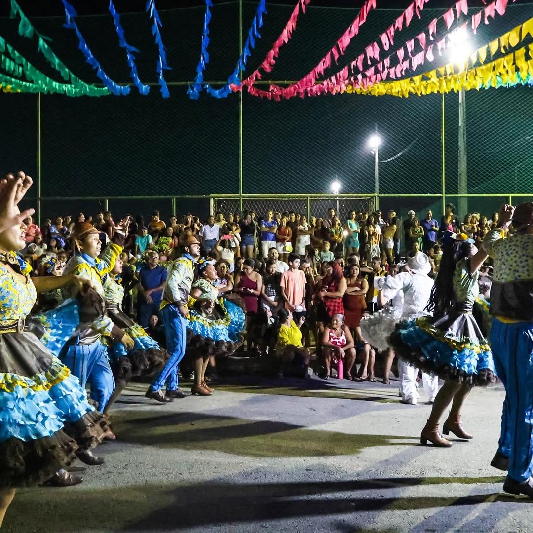Foto noturna de um Arraiá. Dançarinos de quadrilha em trajes típicos coloridos dançam em primeiro plano. O fundo mostra uma grande multidão assistindo, e o alto da cena é decorado com bandeirinhas de festa junina.