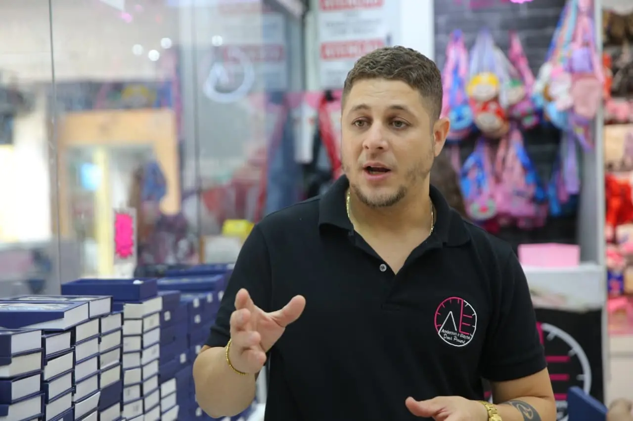 Foto mostra um homem em um ambiente de loja, cercado por várias caixas empilhadas. Ele veste uma camisa polo preta com um logotipo no peito e usa acessórios dourados no pulso e no pescoço. Tem cabelo curto, barba bem aparada e parece estar explicando algo, com a mão levantada em gesto de fala. O fundo é colorido e movimentado, com bolsas e objetos pendurados.