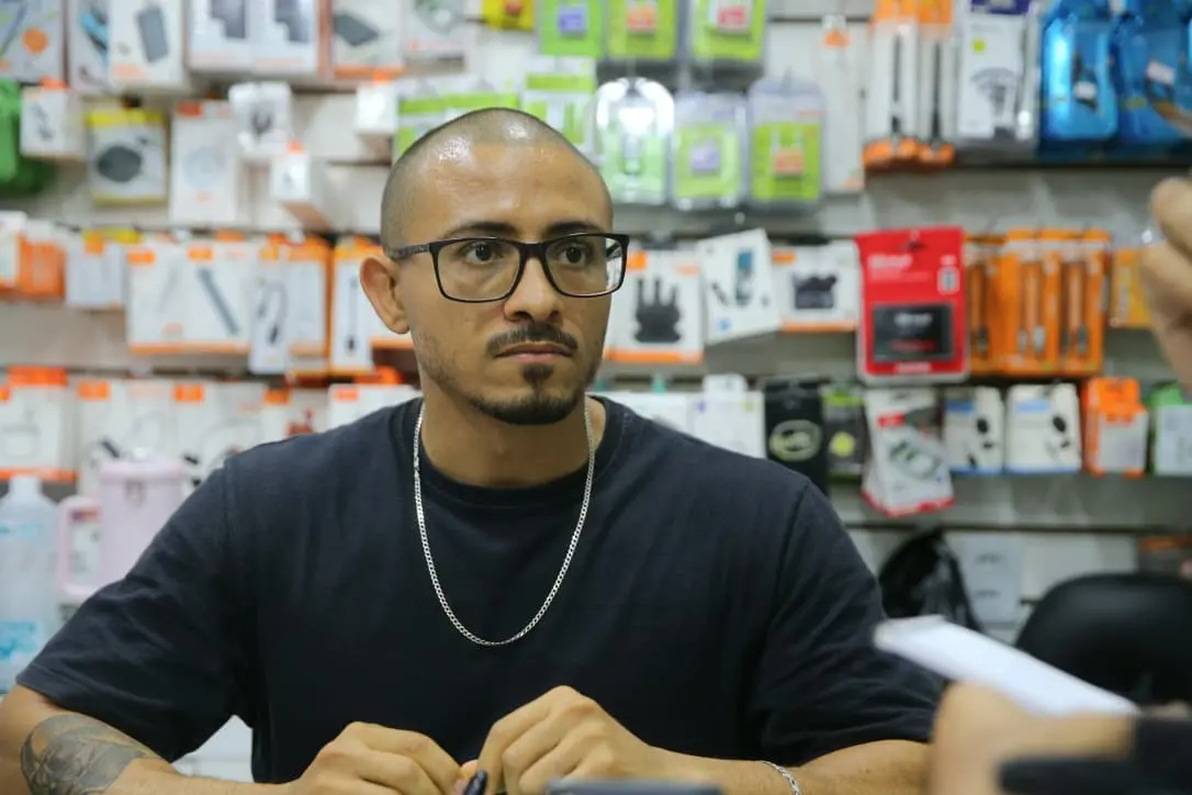 Foto mostra um homem de pé atrás de um balcão em uma loja de eletrônicos e acessórios. Ele é careca, usa óculos de armação preta, corrente prateada e uma camisa preta simples. Seu semblante é sério e atento, possivelmente escutando uma pergunta. Atrás dele há uma parede repleta de produtos embalados, como cabos, carregadores e pequenos eletrônicos.
