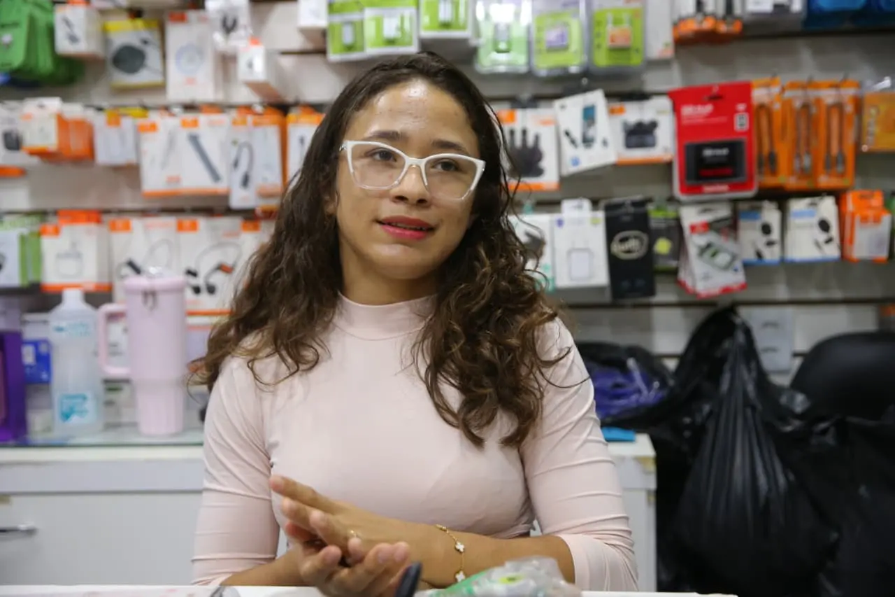 Foto de uma mulher jovem cercada por prateleiras com vários tipos de acessórios eletrônicos. Ela usa óculos brancos de armação grossa, tem cabelo castanho ondulado e veste uma blusa rosa de mangas longas. A expressão é suave e atenta, com as mãos unidas enquanto conversa.