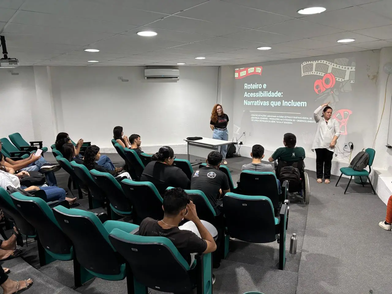 Fotografia de um seminário sobre acessibilidade em cinema. Duas mulheres estão em frente a uma tela que exibe o título 