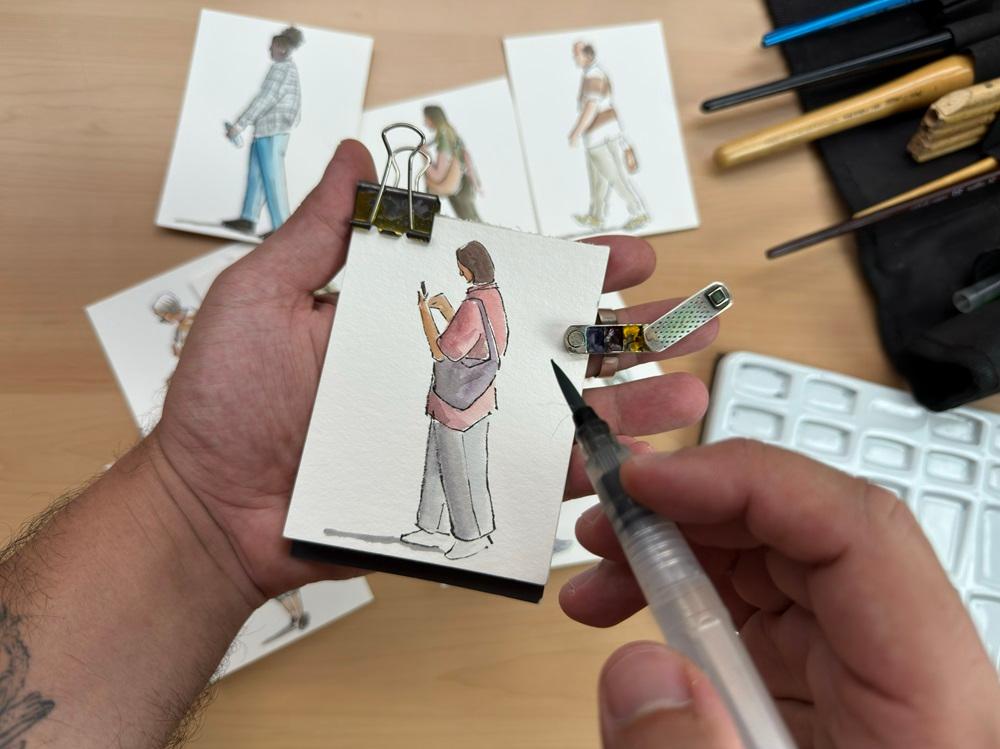 Na imagem, uma foto em primeira pessoa, de cima, mostra uma pessoa segurando um pequeno bloco de notas com um esboço em aquarela de uma figura humana em pé, vestindo uma camisa rosa e calças cinzas, e olhando para o telefone. A mão segura um pincel de água transparente, aplicando tinta. O bloco está preso com um grande clipe de fichário preto. Em primeiro plano e ao fundo, há outros pequenos cartões com esboços em aquarela de figuras humanas em movimento ou paradas. Um estojo de ferramentas e uma paleta de plástico branco estão visíveis à direita. O ambiente é uma superfície de madeira clara.