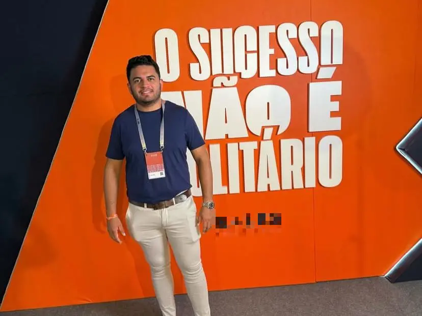 A foto mostra o empresário Iago Viana Nascimento, um homem de cor parda e cabelo preto, que veste uma blusa azul e uma calça bege. Ele usa um crachá. Atrás dele, há uma parede com um letreico, com a frase 'O sucesso não é solitário'.