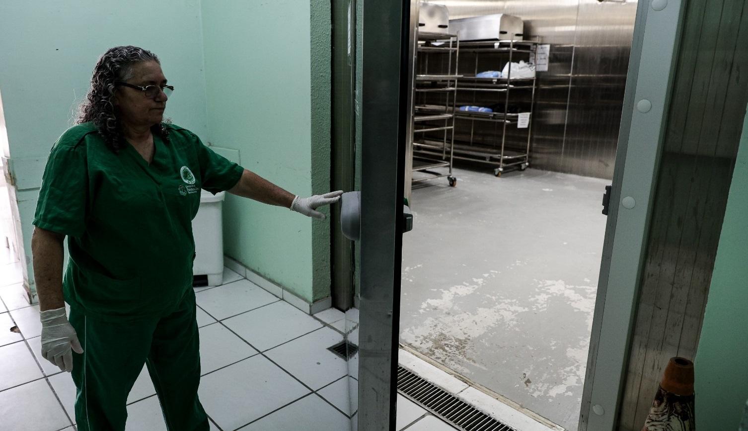 A imagem mostra uma profissional de uniforme verde, usando luvas, abrindo a porta de uma câmara fria no SVO. Atrás da porta, é possível ver um ambiente amplo com piso cimentado e prateleiras metálicas vazias, sugerindo a área de armazenamento refrigerado destinada aos corpos. A foto destaca a rotina silenciosa e protocolar do serviço.