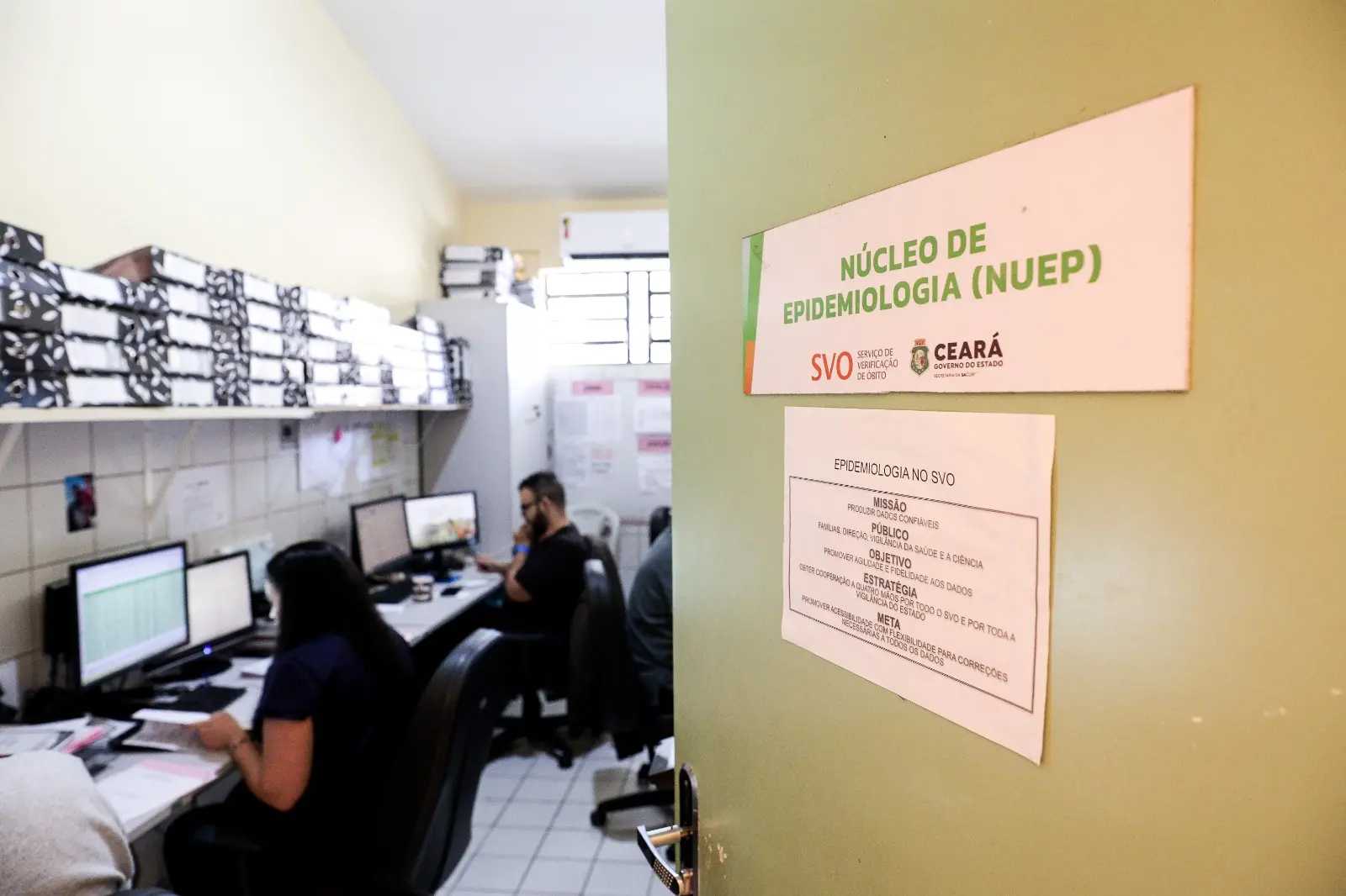 A foto mostra uma sala do Núcleo de Epidemiologia (NUEP) do SVO, com a porta aberta revelando o ambiente interno. Dentro, profissionais trabalham diante de computadores, cercados por pilhas organizadas de arquivos nas prateleiras. A cena transmite rotina técnica e intensa de análise de dados, em um espaço pequeno e funcional.