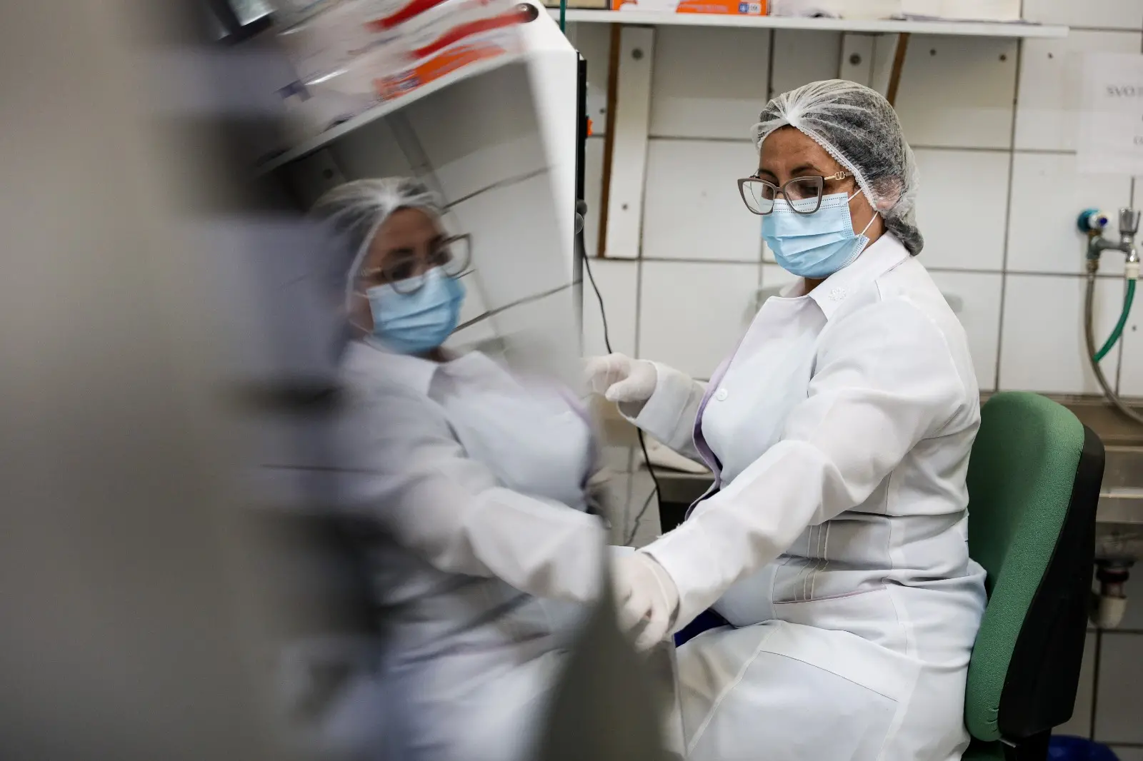 Uma técnica de laboratório, vestida com jaleco branco, touca e máscara cirúrgica, e óculos, está sentada realizando uma análise. A imagem é refletida em uma superfície brilhante à esquerda, enquanto a profissional se concentra em sua tarefa.